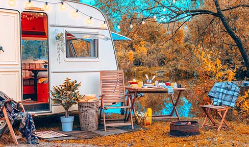 A caravan parked in a wooded area, accompanied by a table and chairs set up nearby for outdoor relaxation